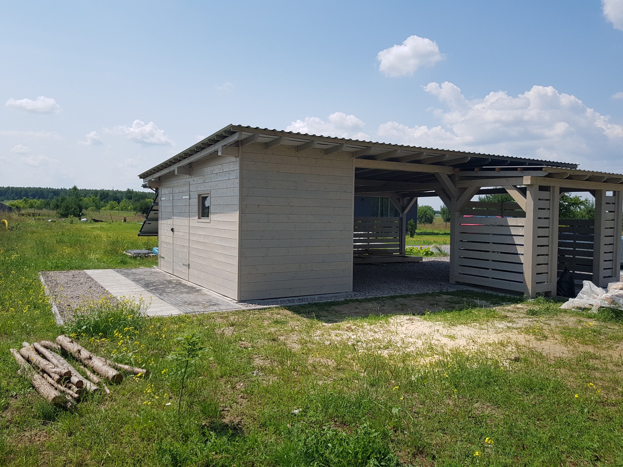Drewniana konstrukcja składająca się z zamkniętej szopy z dwuskrzydłowymi drzwiami i otwartej zadaszonej przestrzeni, obie w jasnoszarym kolorze, na tle zielonej łąki z żółtymi kwiatami...
