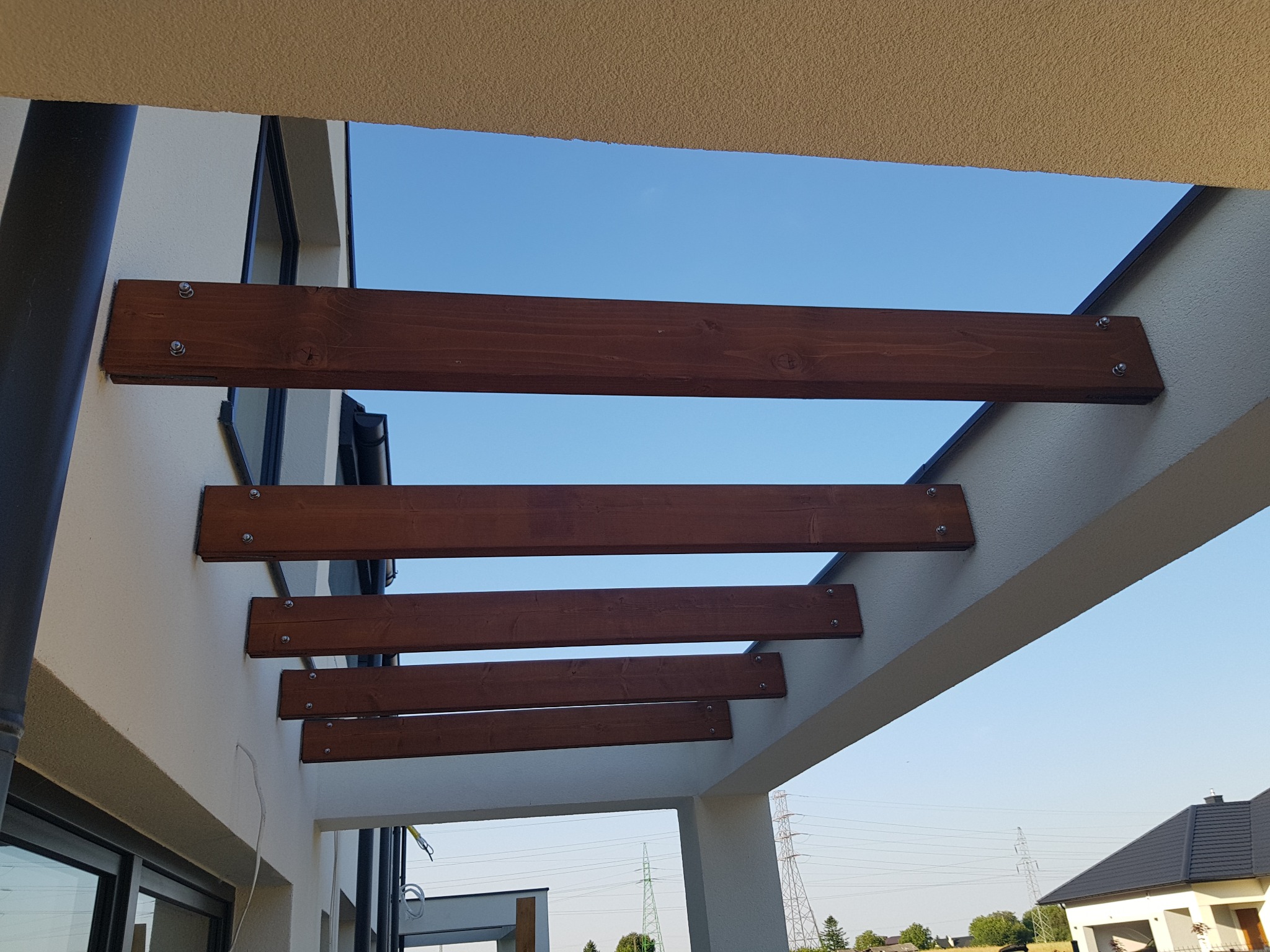 Drewniana konstrukcja z belek przymocowanych do ściany budynku, widok z dołu na błękitne niebo, elementy mocujące widoczne.