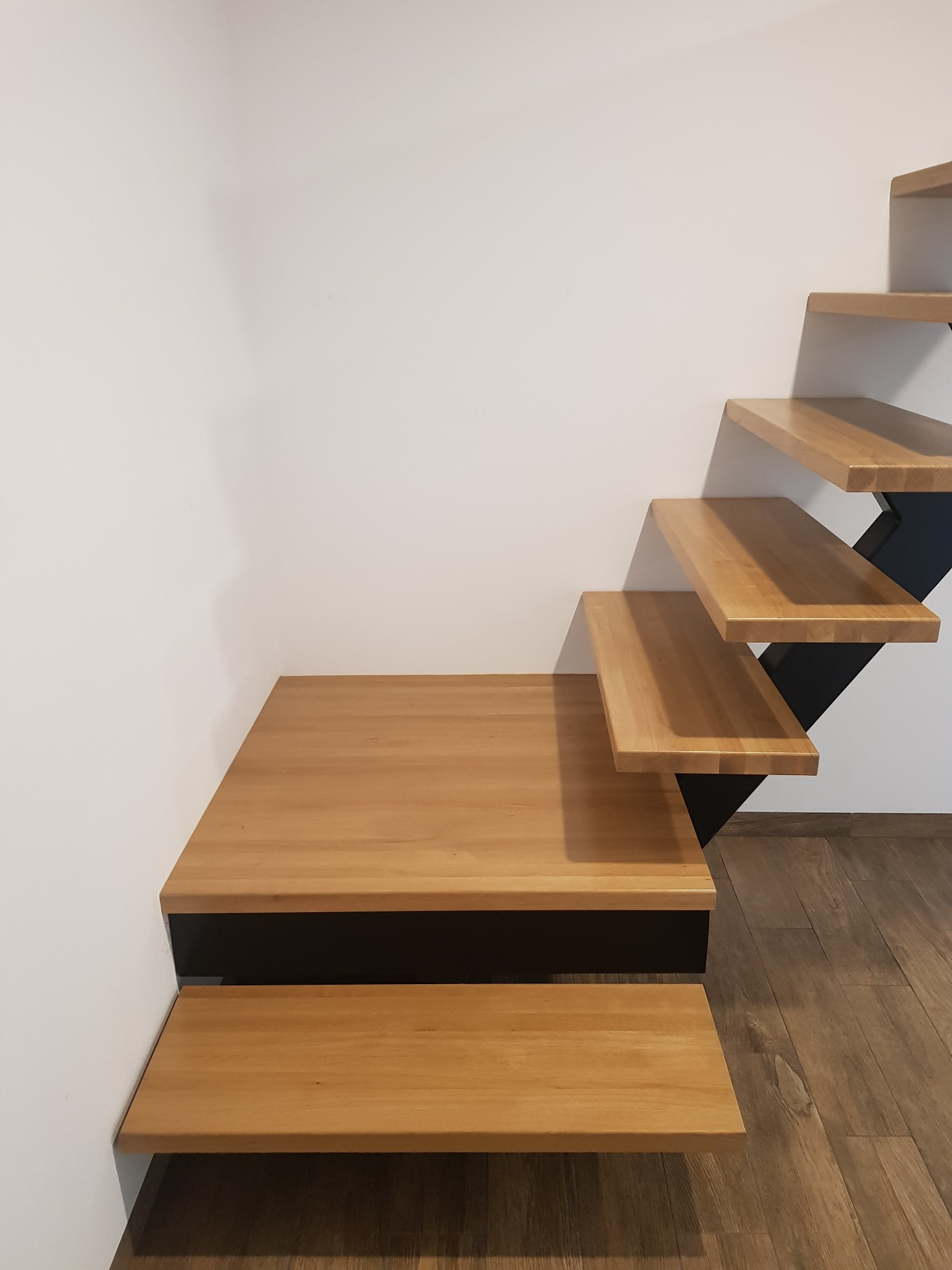 Nowoczesne schody drewniane z prostymi stopniami i czarną, metalową konstrukcją wsporczą, przylegające do białej ściany, widoczne z dołu.