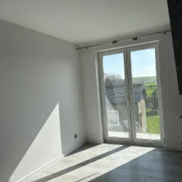 Pusty, jasny pokój z białymi ścianami, szarą podłogą imitującą panele i dużym oknem z widokiem na budynek gospodarczy i zielone wzgórza w tle.