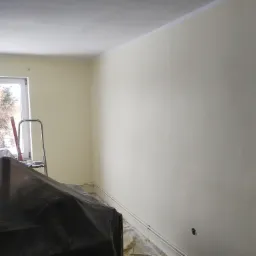 Świeżo pomalowane ściany w pokoju, zabezpieczone meble folią ochronną, drabina przy oknie, widoczne listwy przypodłogowe i gniazdko elektryczne.