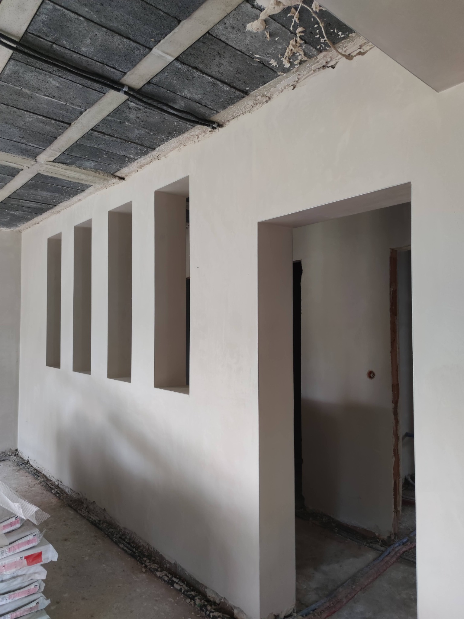 Wnętrze po nałożeniu gładzi gipsowej, widoczne pionowe wnęki w ścianie i surowy betonowy strop z instalacją elektryczną. W tle otwór drzwiowy.