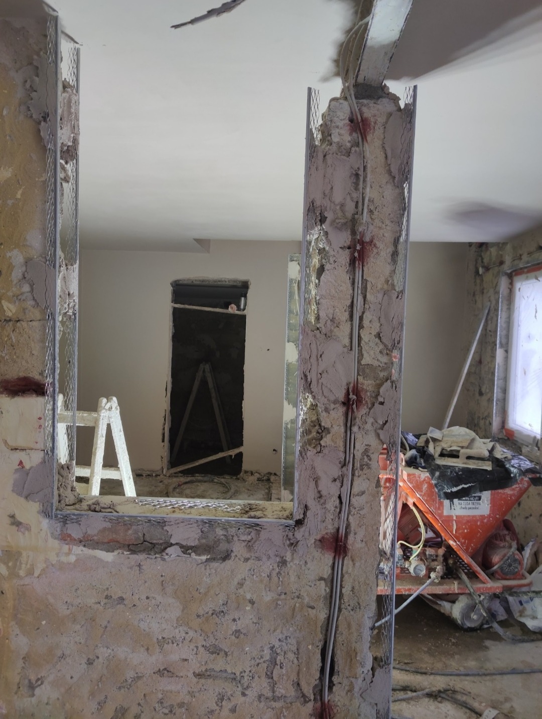 Przygotowanie ściany do nałożenia tynku maszynowego, widoczne profile narożnikowe z siatką i przewody elektryczne przymocowane do ściany, w tle maszyna do tynkowania.