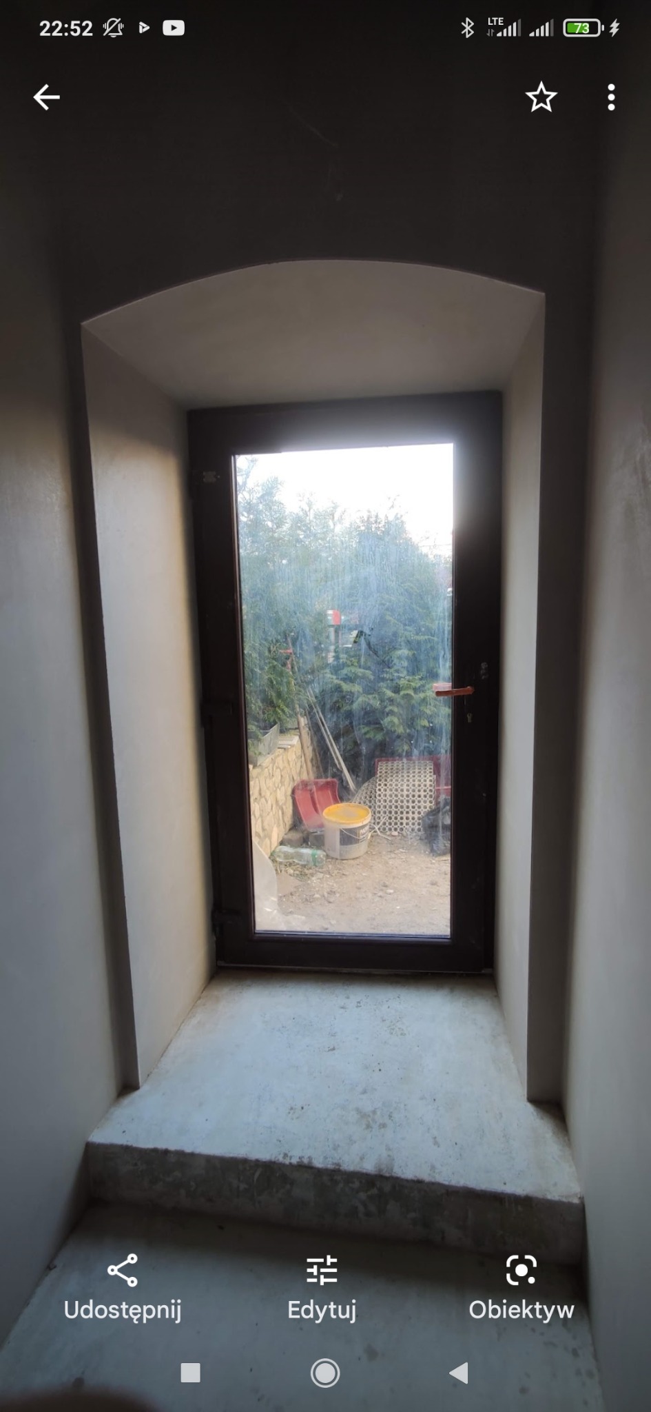 Świeżo otynkowane maszynowo ściany w Częstochowie, widoczne przez otwarte drzwi na zewnątrz, gdzie znajduje się ogród i narzędzia budowlane.