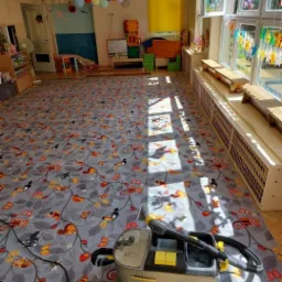 Karcher Professional na wzorzystym dywanie w przedszkolu, widoczne zabawki i dekoracje, promienie słońca wpadają przez okno.