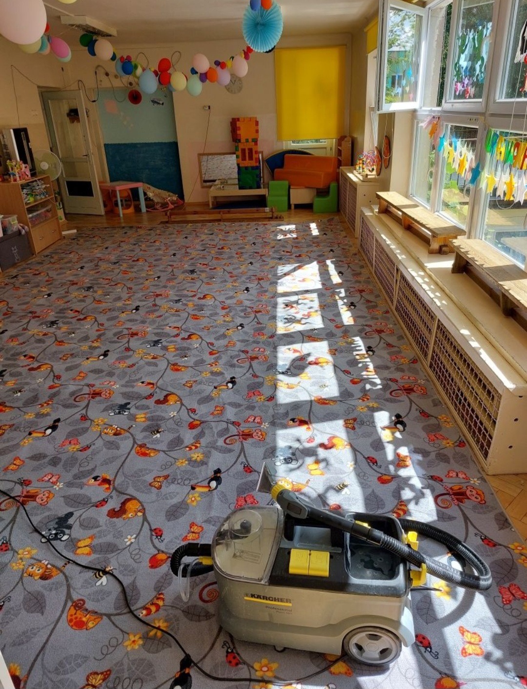 Karcher Professional na wzorzystym dywanie w przedszkolu, widoczne zabawki i dekoracje, promienie słońca wpadają przez okno.