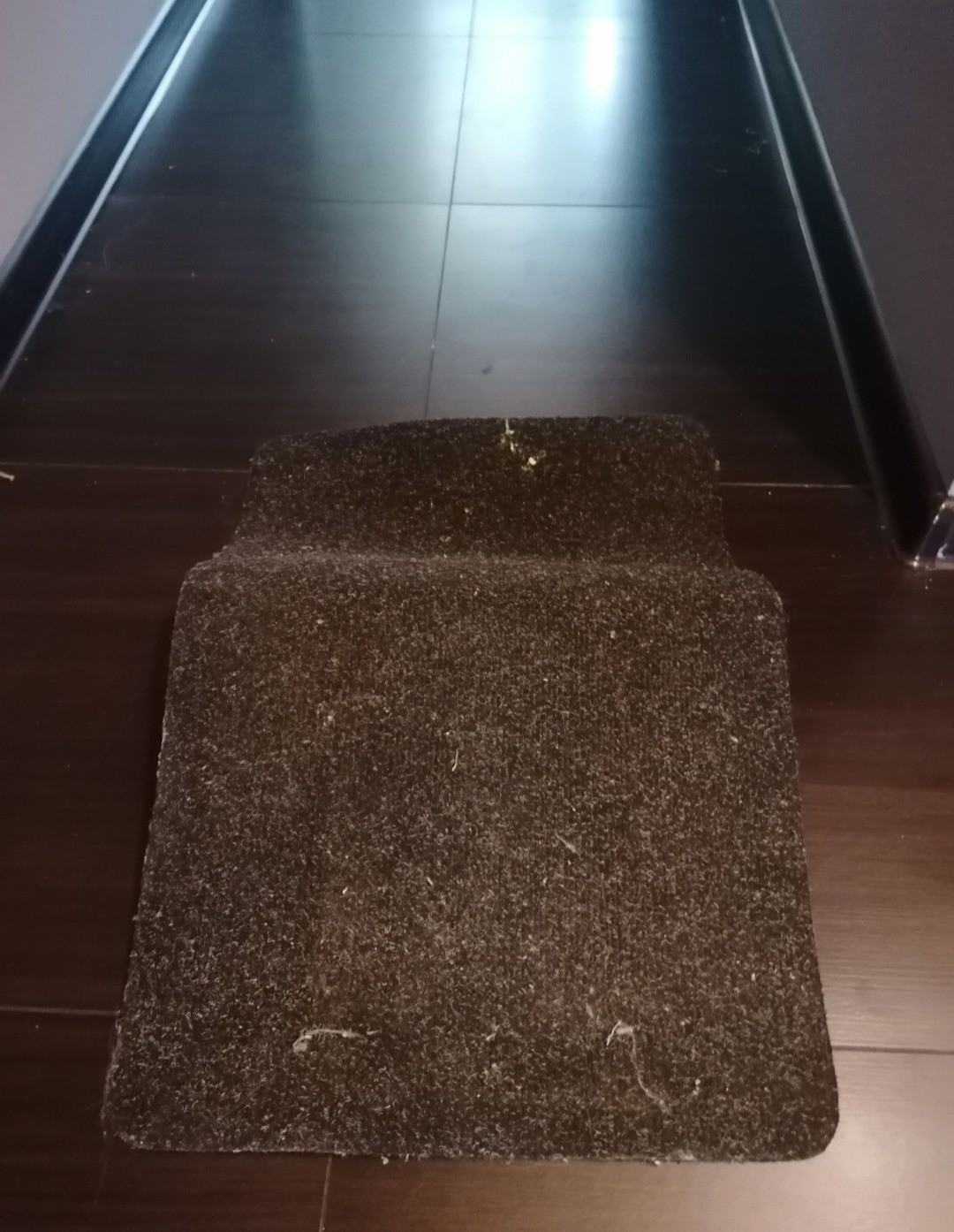 Ciemny, złożony dywanik z widocznymi śladami użytkowania i drobnymi zabrudzeniami, leżący na ciemnej, lakierowanej podłodze.