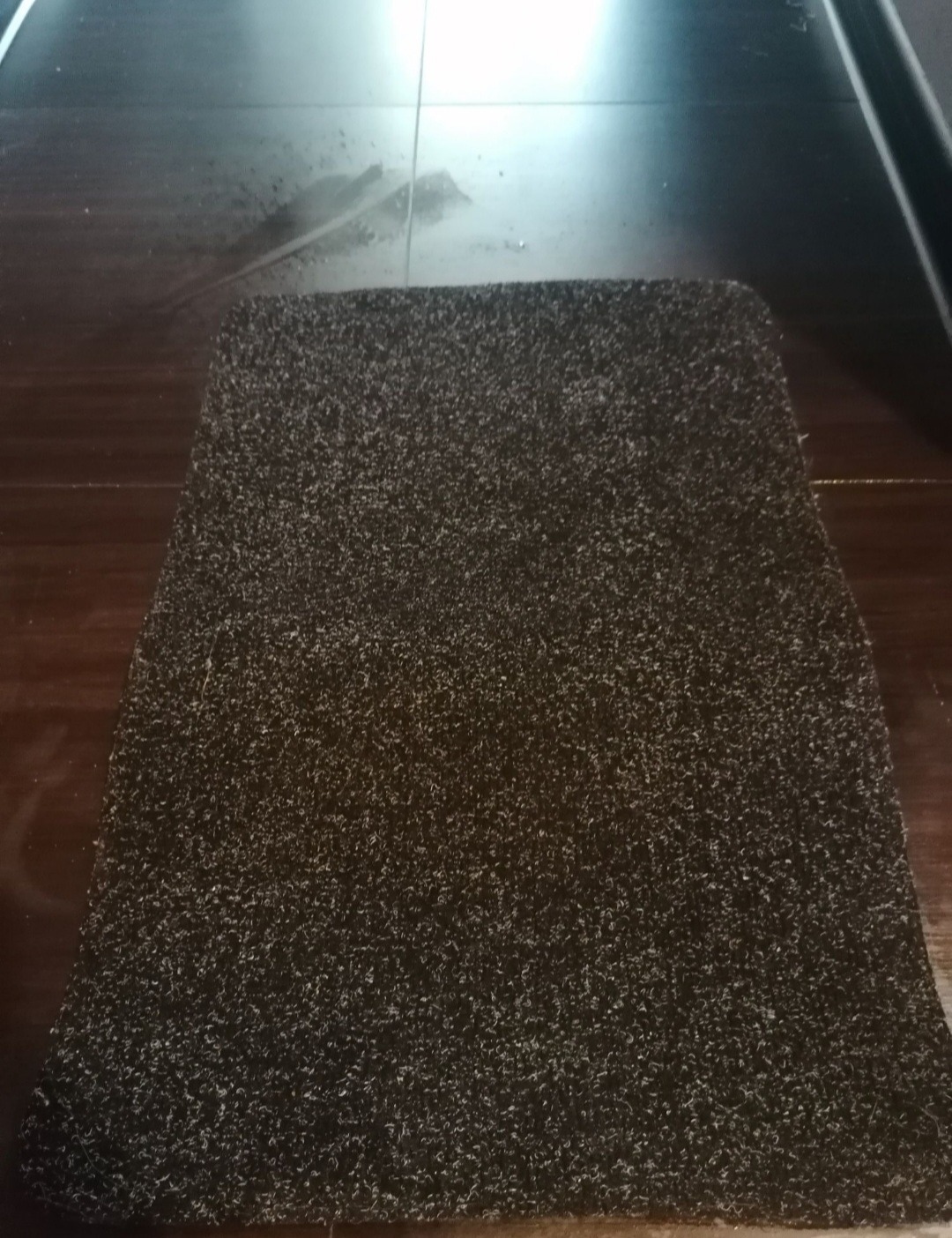 Ciemny dywanik z krótkim włosiem leżący na błyszczącej, ciemnej podłodze z widocznymi plamami wody.