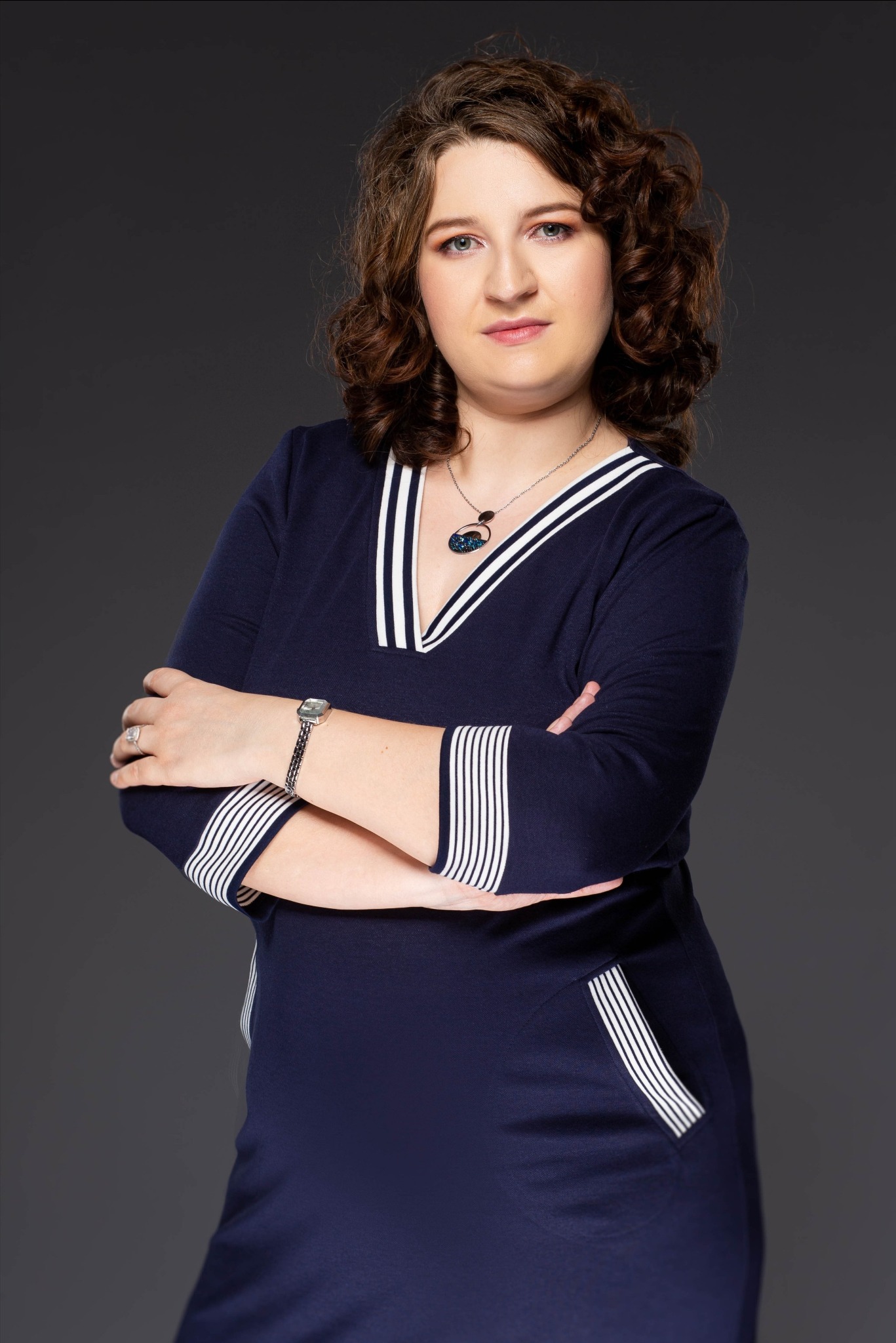 Portret kobiety z kręconymi włosami, ubranej w granatową sukienkę z białymi paskami, z rękami skrzyżowanymi na piersiach, na szarym tle.