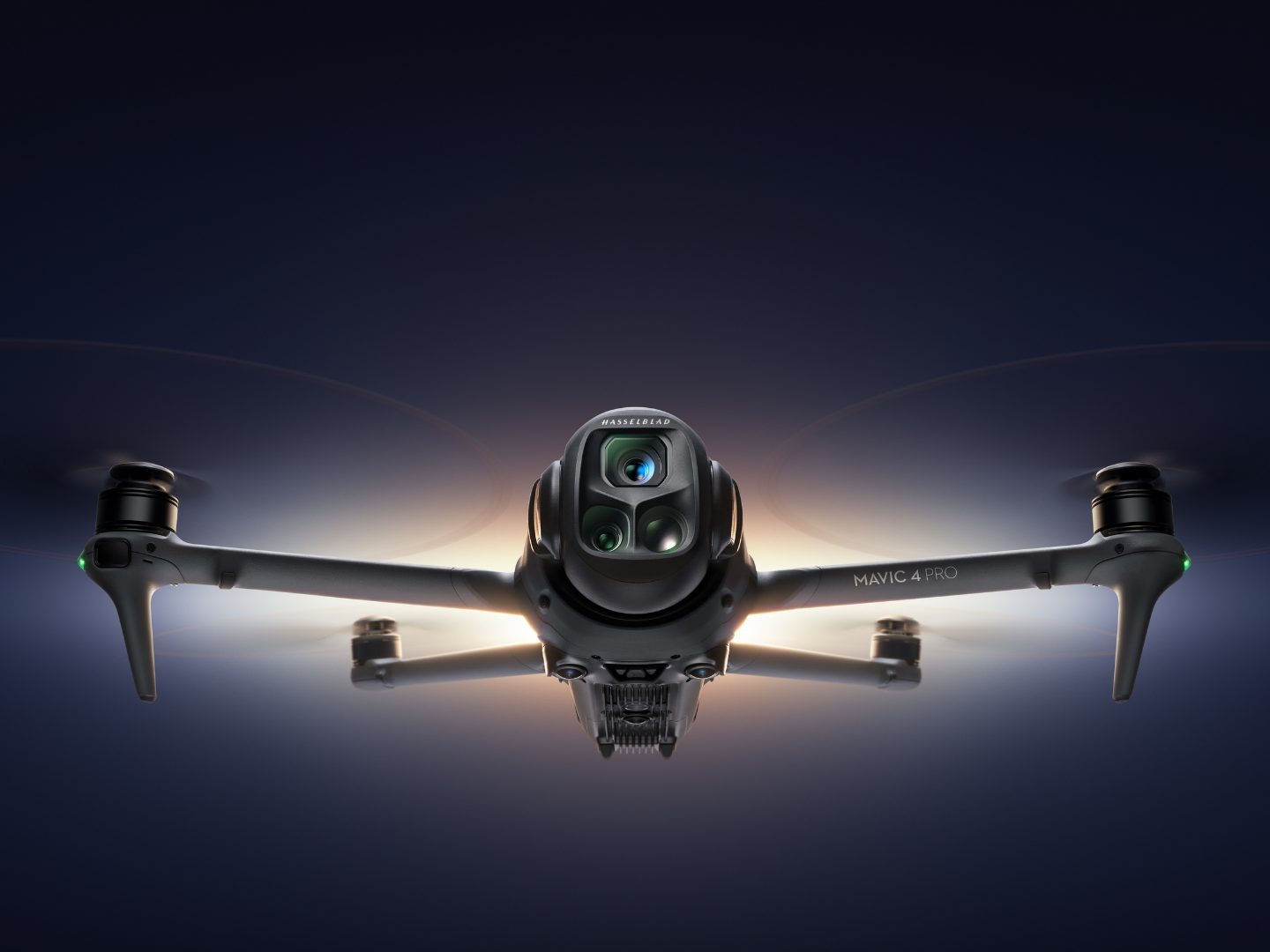 Czarny dron Mavic 4 Pro z kamerą Hasselblad w locie, oświetlony od dołu, na ciemnym tle. Widoczne wirniki i diody sygnalizacyjne.