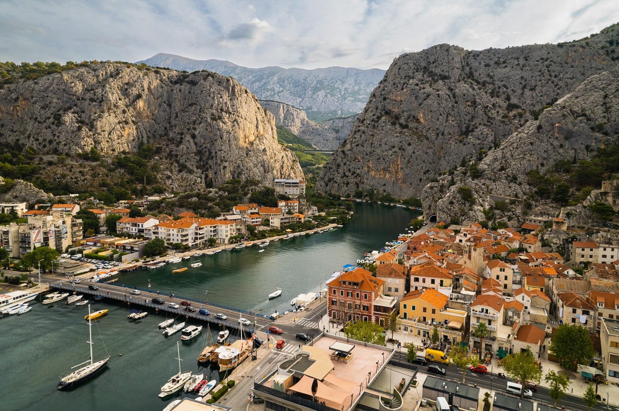 Panoramiczny widok na miasto z rzeką, mostem, łodziami i górami w tle, wykonany z góry. Charakterystyczna architektura z czerwonymi dachami.
