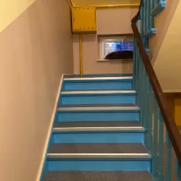 Schody w bloku pomalowane na niebiesko z szarymi nakładkami na stopniach, ściany w kolorze żółtym i różowym, żółta skrzynka elektryczna i okno w tle.