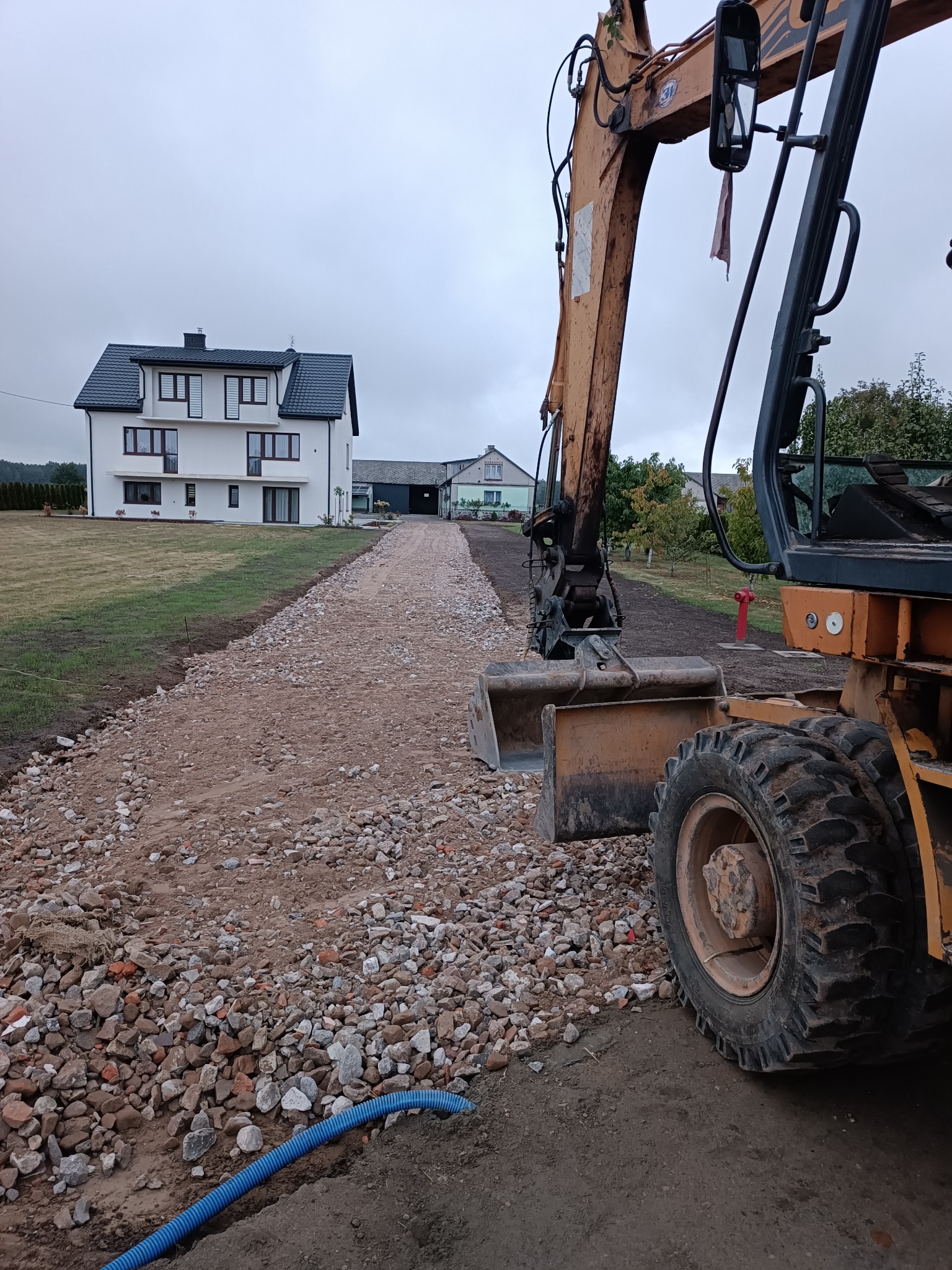 Żółta koparka kołowa na żwirowej drodze w trakcie budowy, w tle biały dom z ciemnym dachem i budynki gospodarcze, widoczny niebieski drenarski wąż.
