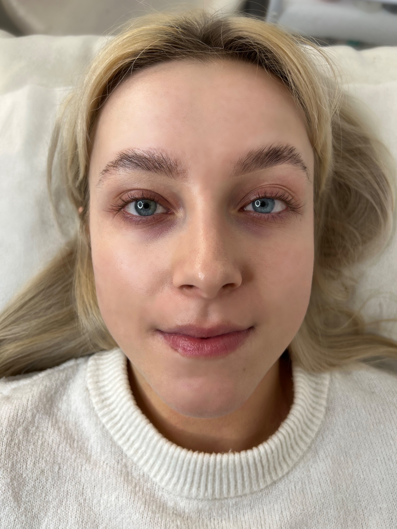 Blondwłosa kobieta z widocznymi brwiami po zabiegu laminacji, leżąca na białym podłożu, z delikatnym makijażem.