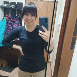 Uśmiechnięta kobieta robi selfie w lustrze w przedpokoju; widoczna półka z ubraniami i butami w tle.