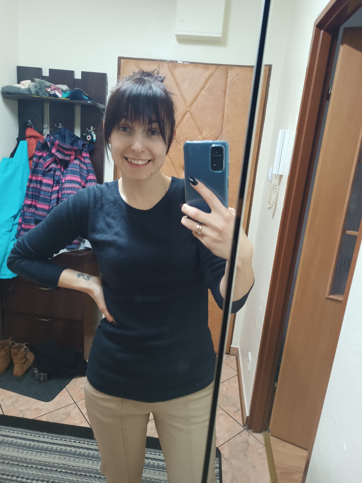 Uśmiechnięta kobieta robi selfie w lustrze w przedpokoju; widoczna półka z ubraniami i butami w tle.