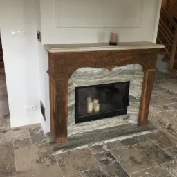 Elegancki kominek z marmurową obudową i rzeźbionym drewnianym portalem, ustawiony na podłodze wyłożonej nieregularnymi płytkami w odcieniach beżu i brązu; w tle widoczne drewniane schody i fragment...