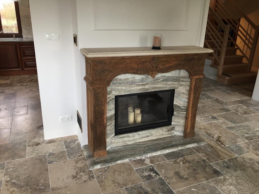 Elegancki kominek z marmurową obudową i rzeźbionym drewnianym portalem, ustawiony na podłodze wyłożonej nieregularnymi płytkami w odcieniach beżu i brązu; w tle widoczne drewniane schody i fragment...