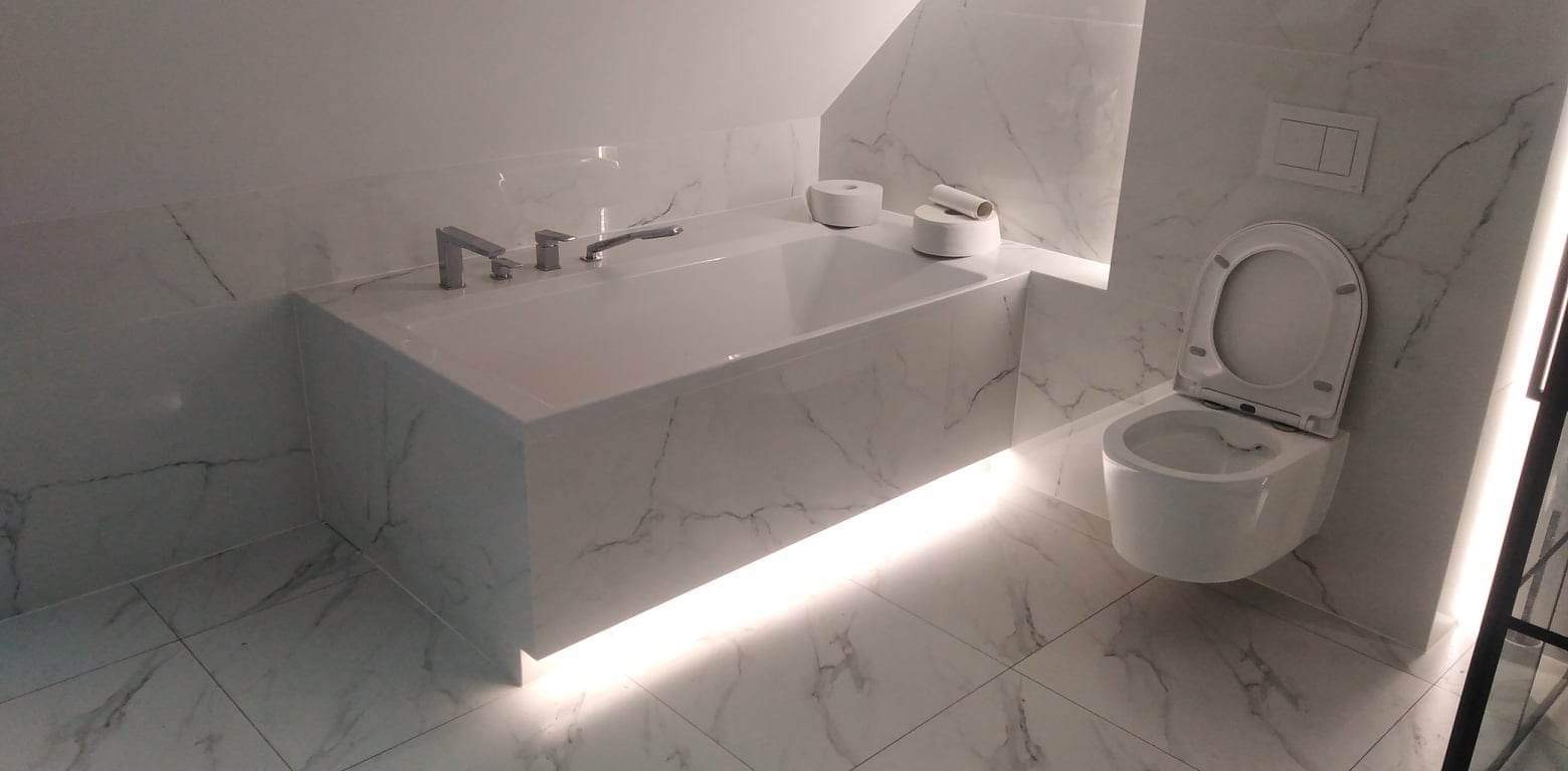 Nowoczesna łazienka w bieli z marmurowym wzorem, podświetlana wanna obudowana płytami, wisząca toaleta i minimalistyczna armatura.