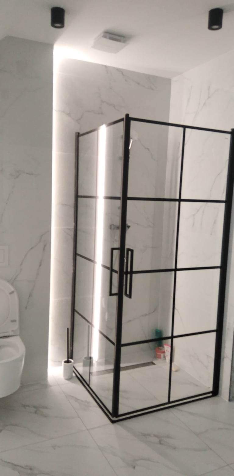 Nowoczesna kabina prysznicowa z czarnymi profilami i szklanymi ścianami w łazience wykończonej marmurowymi płytkami, widoczna toaleta z lewej strony, oświetlenie LED w pionowym panelu.