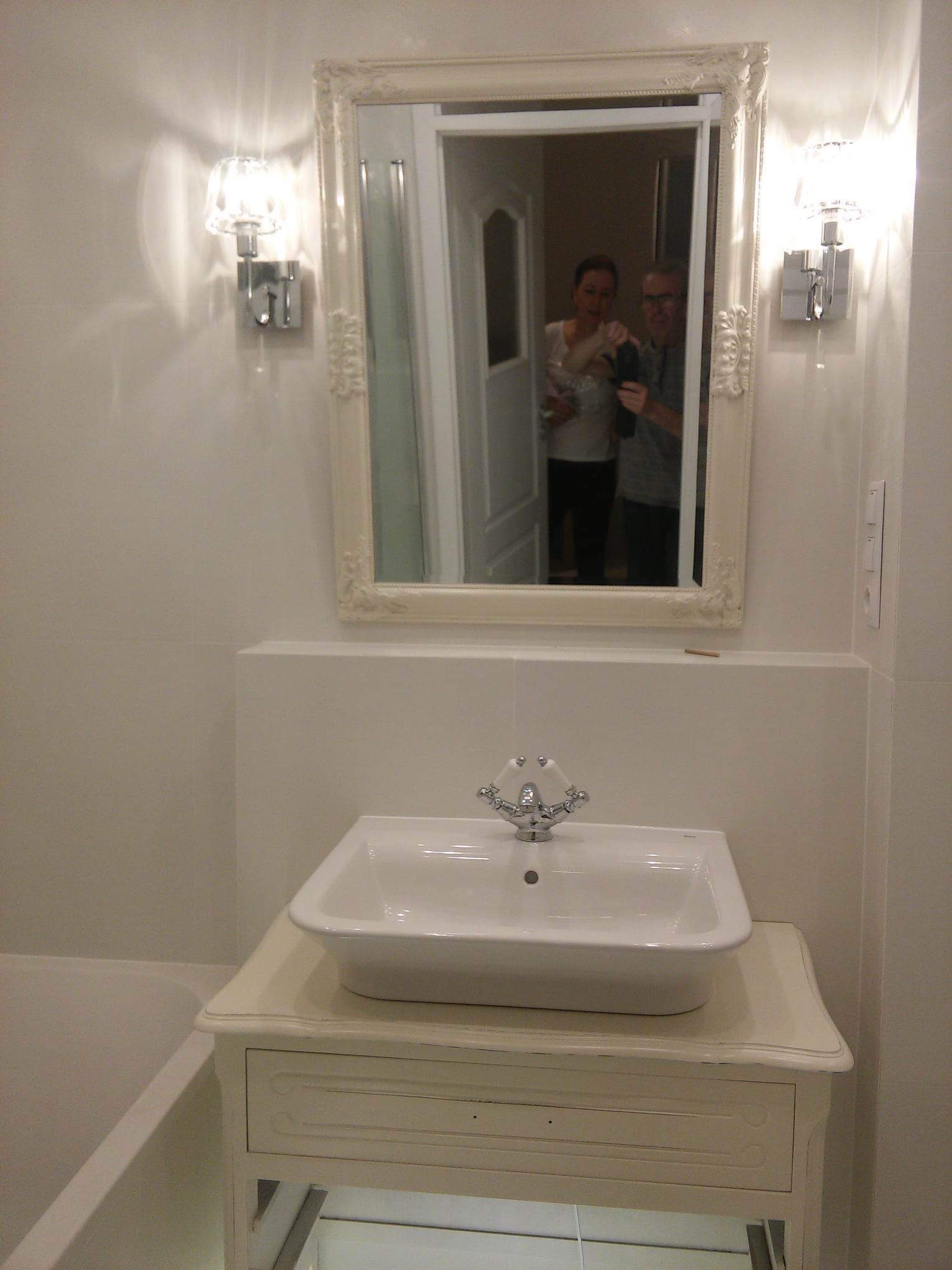 Biała umywalka nablatowa z ozdobną baterią, umieszczona na stylizowanej szafce z szufladą w jasnej łazience, z widocznym w lustrze odbiciem osób robiących zdjęcie.
