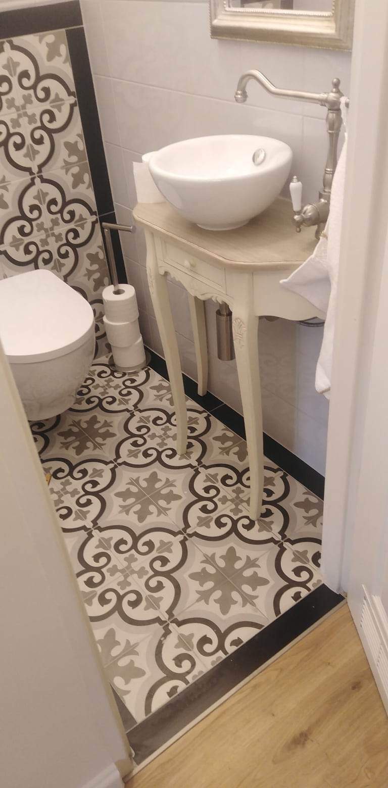 Ukończona łazienka z wzorzystą, szarą i białą glazurą podłogową, toaleta i umywalka na dekoracyjnej, kremowej konsoli z armaturą w stylu retro.