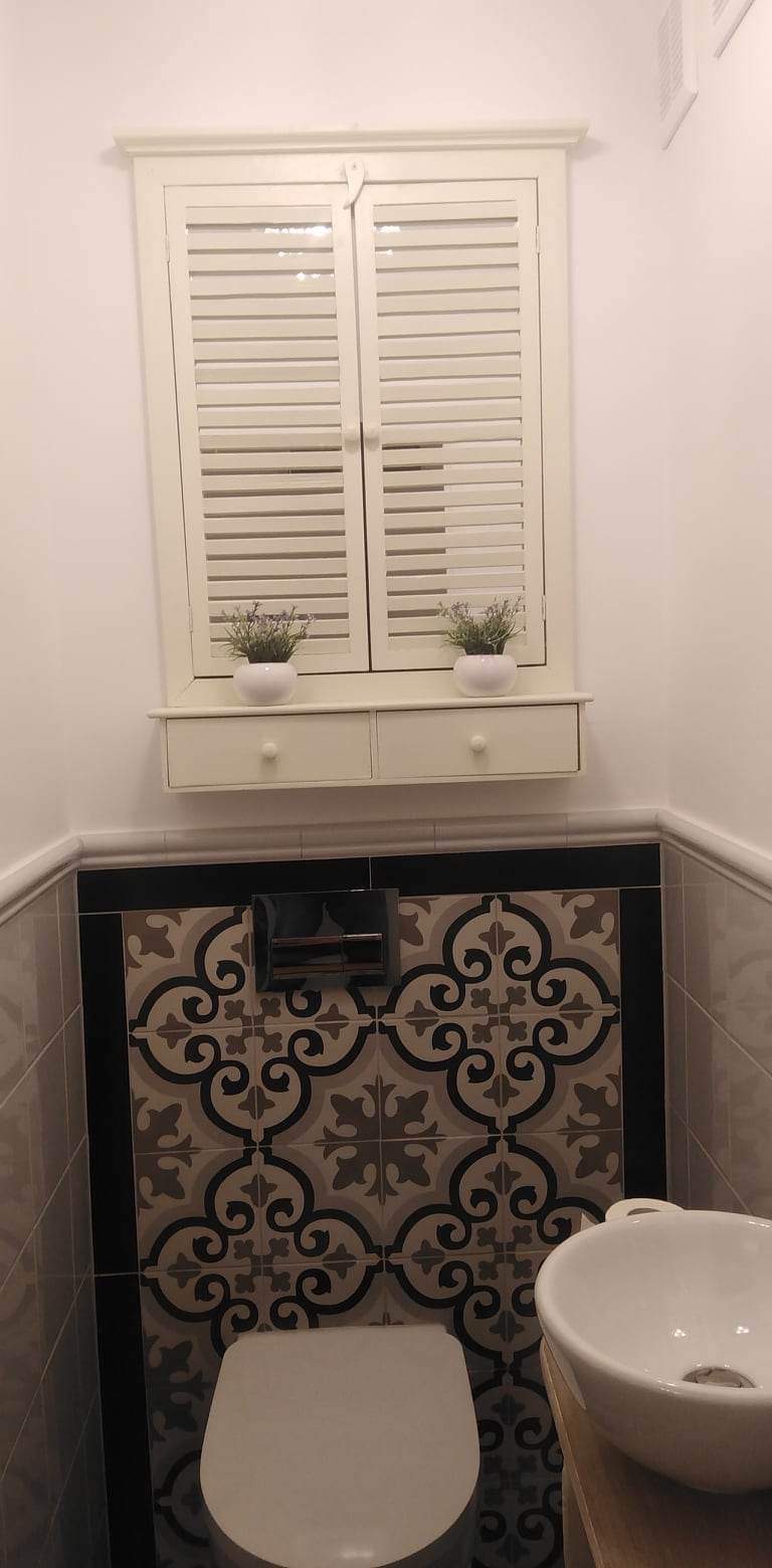 Wykończona łazienka z dekoracyjnymi płytkami w odcieniach szarości i beżu, białą toaletą oraz umywalką. Nad toaletą widoczna zabudowa z żaluzjowymi drzwiczkami i małymi doniczkami z kwiatami.