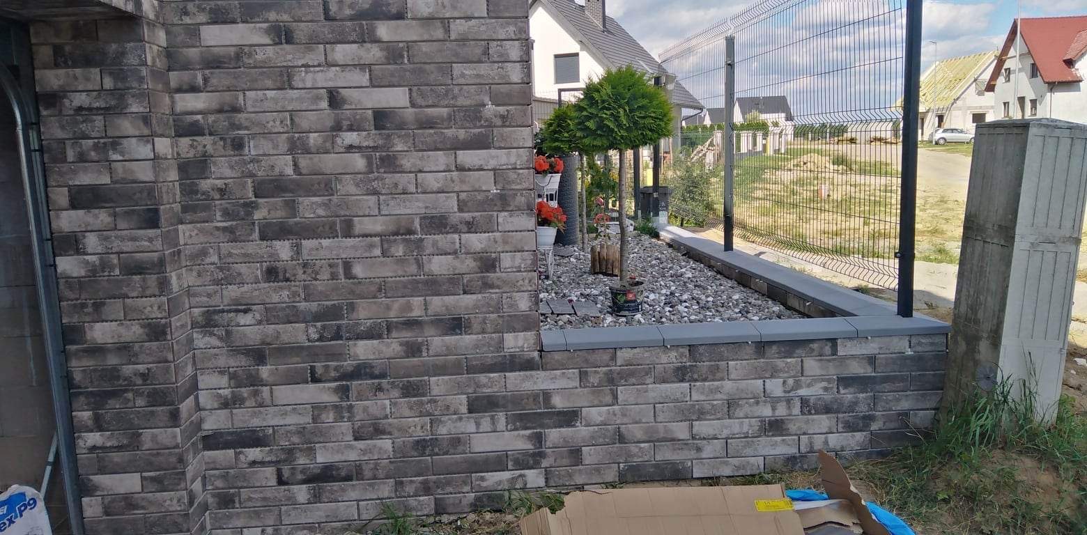 Murowana ściana z szarych cegieł, stanowiąca element małej architektury ogrodowej, z widocznymi roślinami i ogrodzeniem panelowym w tle.
