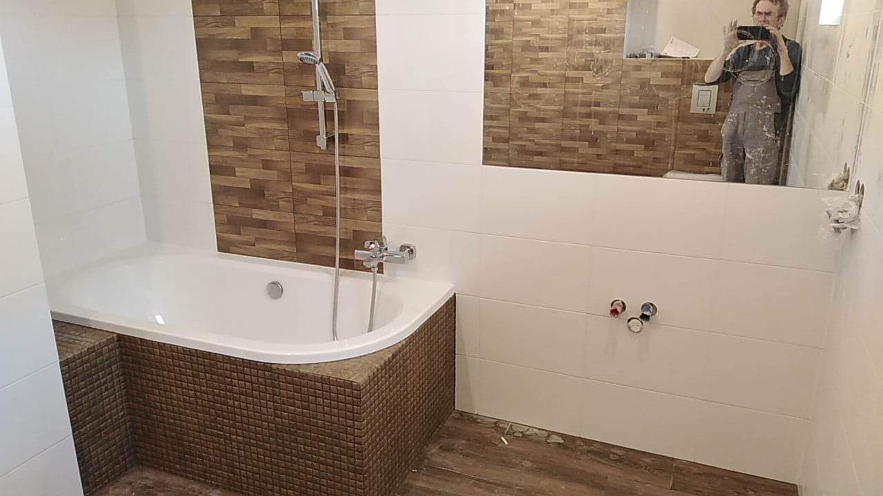 Nowoczesna łazienka w trakcie wykańczania: białe płytki ścienne, mozaika na obudowie wanny, drewnopodobne płytki na ścianie i podłodze, widoczne przyłącza wodne, odbicie fotografa w lustrze.