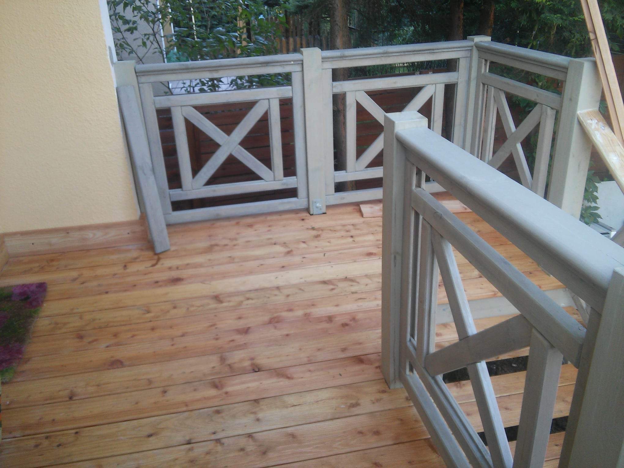 Drewniany taras z pomalowaną na szaro balustradą z motywem krzyży, widok z góry, fragment ściany budynku w tle.