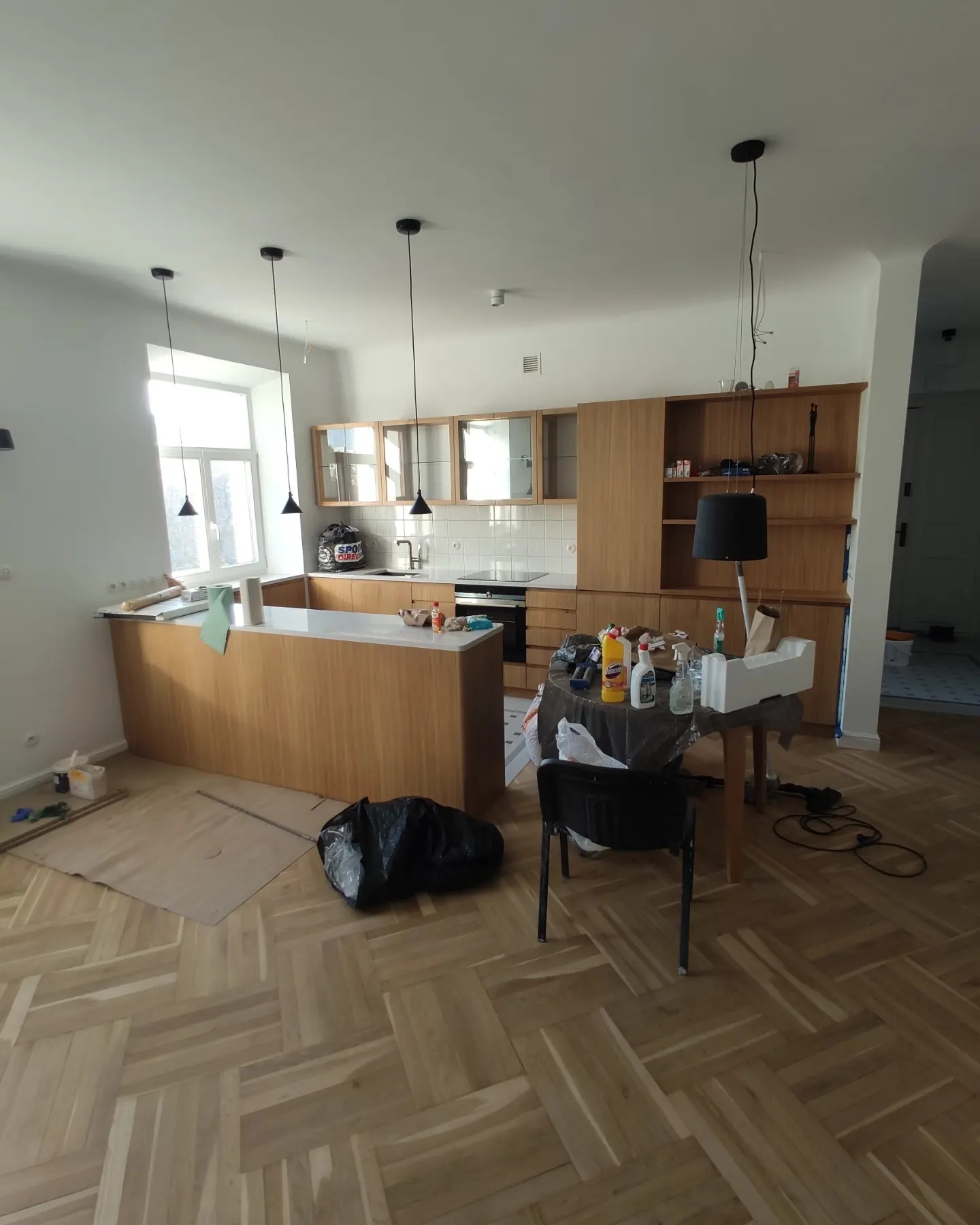 Kuchnia w trakcie prac wykończeniowych: drewniane meble kuchenne z białym blatem, podłoga w jodełkę, czarne lampy wiszące, stół roboczy z chemią budowlaną i narzędziami, zabezpieczona podłoga.