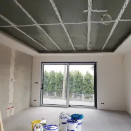 Wnętrze w Żarach w trakcie prac wykończeniowych: sufit z płyt kartonowo-gipsowych na ruszcie, zabezpieczony folią i taśmą, przygotowany do dalszych prac. Widoczne puszki elektryczne w ścianie...