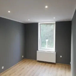 Pokój po remoncie: szare ściany, jasna podłoga imitująca drewno, białe okno z grzejnikiem, w suficie zamontowane halogenowe lampy.