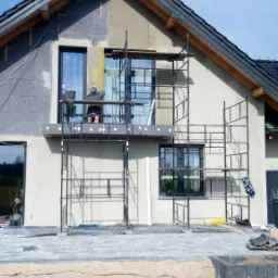 Prace na elewacji domu jednorodzinnego: ocieplanie ścian zewnętrznych styropianem, widoczne rusztowania i pracownik na wysokości, teren wokół domu w trakcie zagospodarowywania.