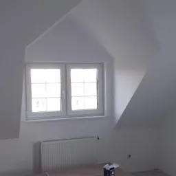 Poddasze ze świeżo pomalowanymi na biało ścianami, okno z białymi ramami, grzejnik oraz stos kartonów z panelami podłogowymi marki Ikerra.