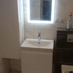 Nowoczesna łazienka z białymi marmurkowymi płytkami, podświetlanym lustrem nad białą umywalką z szafką, oraz bidetem w rogu.