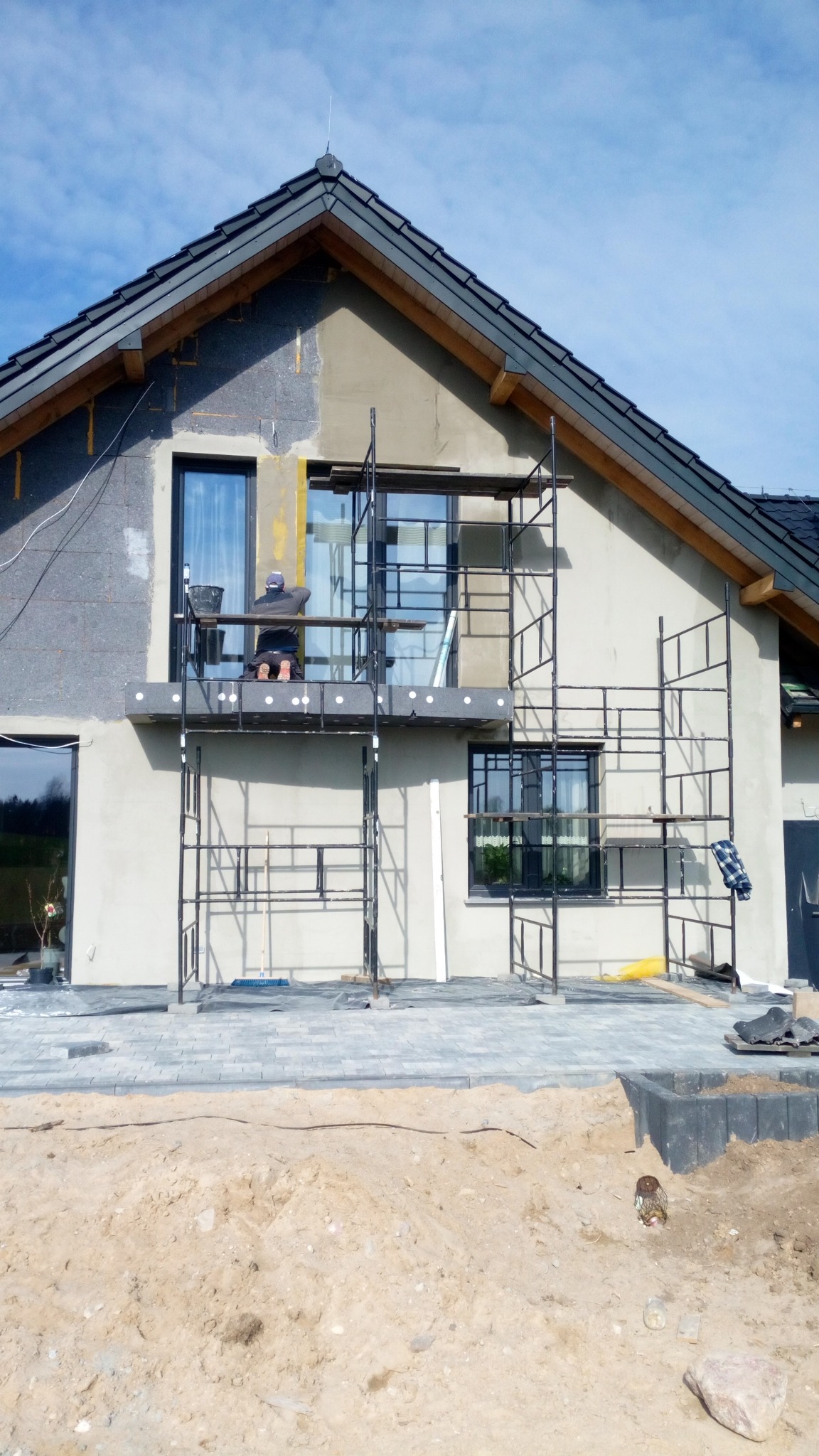 Prace na elewacji domu jednorodzinnego: ocieplanie ścian zewnętrznych styropianem, widoczne rusztowania i pracownik na wysokości, teren wokół domu w trakcie zagospodarowywania.