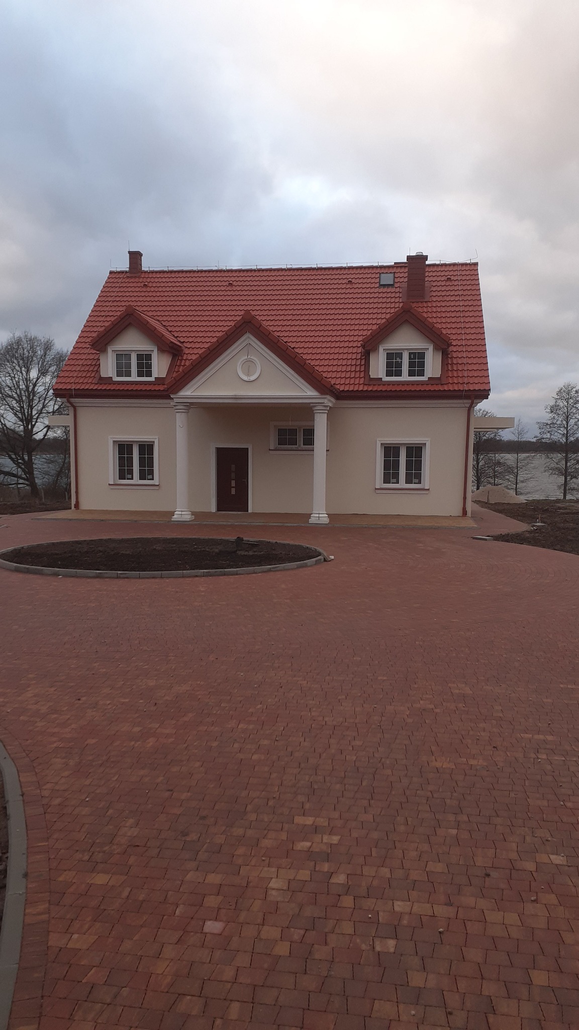 Nowo wybudowany dom jednorodzinny z czerwoną dachówką, jasną elewacją i kolumnami przy wejściu, otoczony brukowanym podjazdem i przygotowanym miejscem na trawnik, widok z przodu w pochmurny dzień.