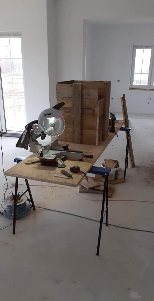 Wnętrze w trakcie wykończenia: drewniana konstrukcja skrzyniowa na stole warsztatowym z piłą tarczową Metabo, w tle surowe ściany i okna.