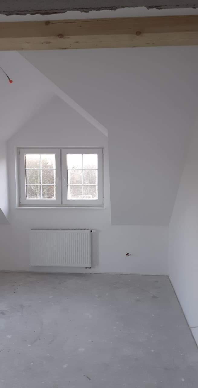 Poddasze w stanie surowym z oknem, grzejnikiem i drewnianą belką stropową, świeżo pomalowane na biało.