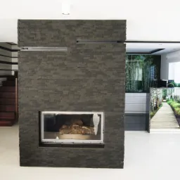 Nowoczesny salon z kominkiem obłożonym szarą, kamienną okładziną, widoczne schody z drewnianymi stopniami i fototapeta z motywem leśnej ścieżki w tle.