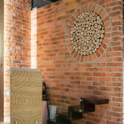 Ceglana ściana z okrągłą dekoracją z drewnianych krążków, obok drewniany panel dekoracyjny z koncentrycznymi okręgami i półkami w formie schodków.