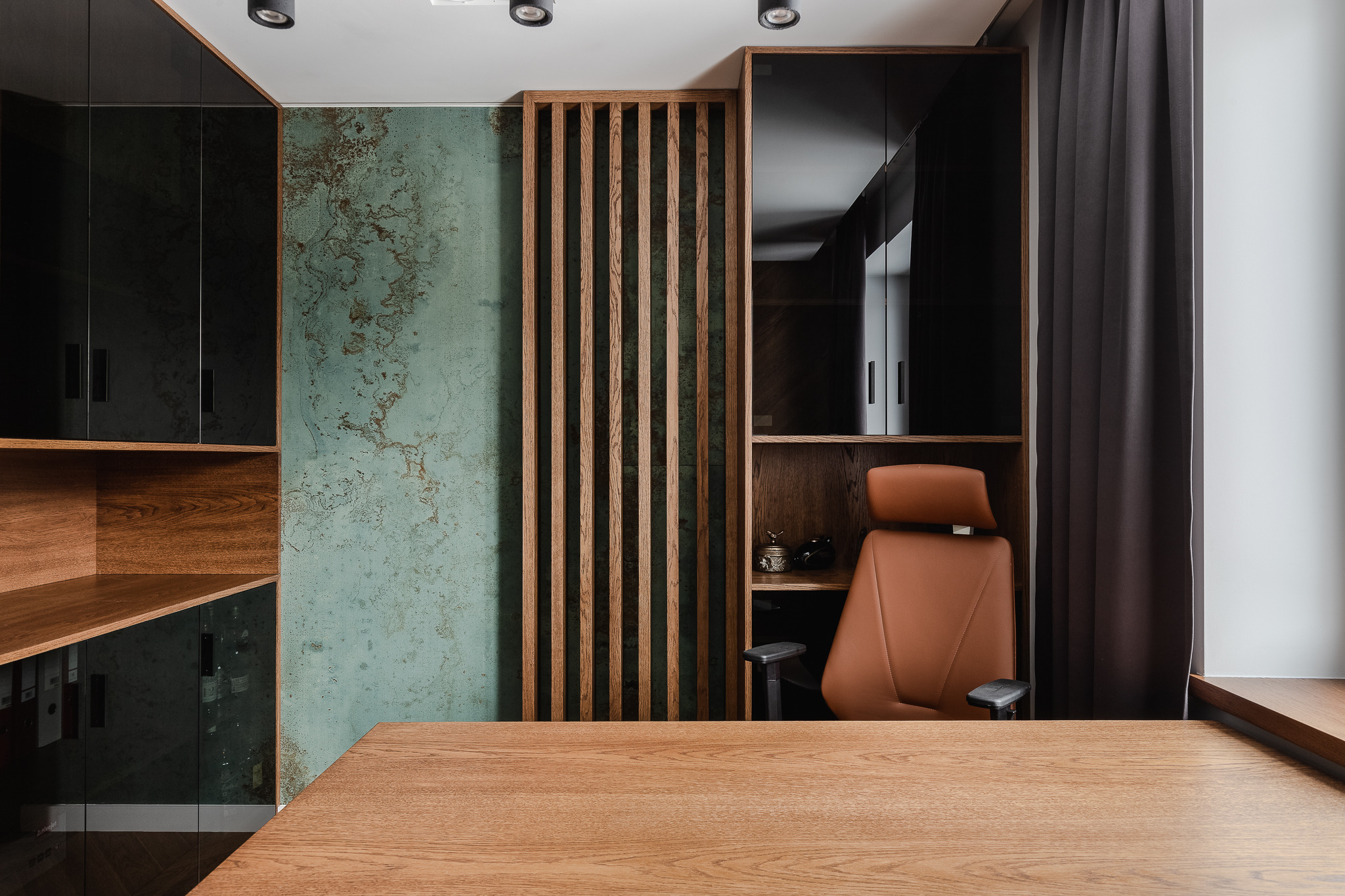 Nowoczesne biuro z drewnianym biurkiem, ścianą w odcieniach turkusu i brązu, oraz krzesłem biurowym z brązowej skóry ekologicznej. Na półkach widoczne akcesoria biurowe i czarne, lakierowane szafki.