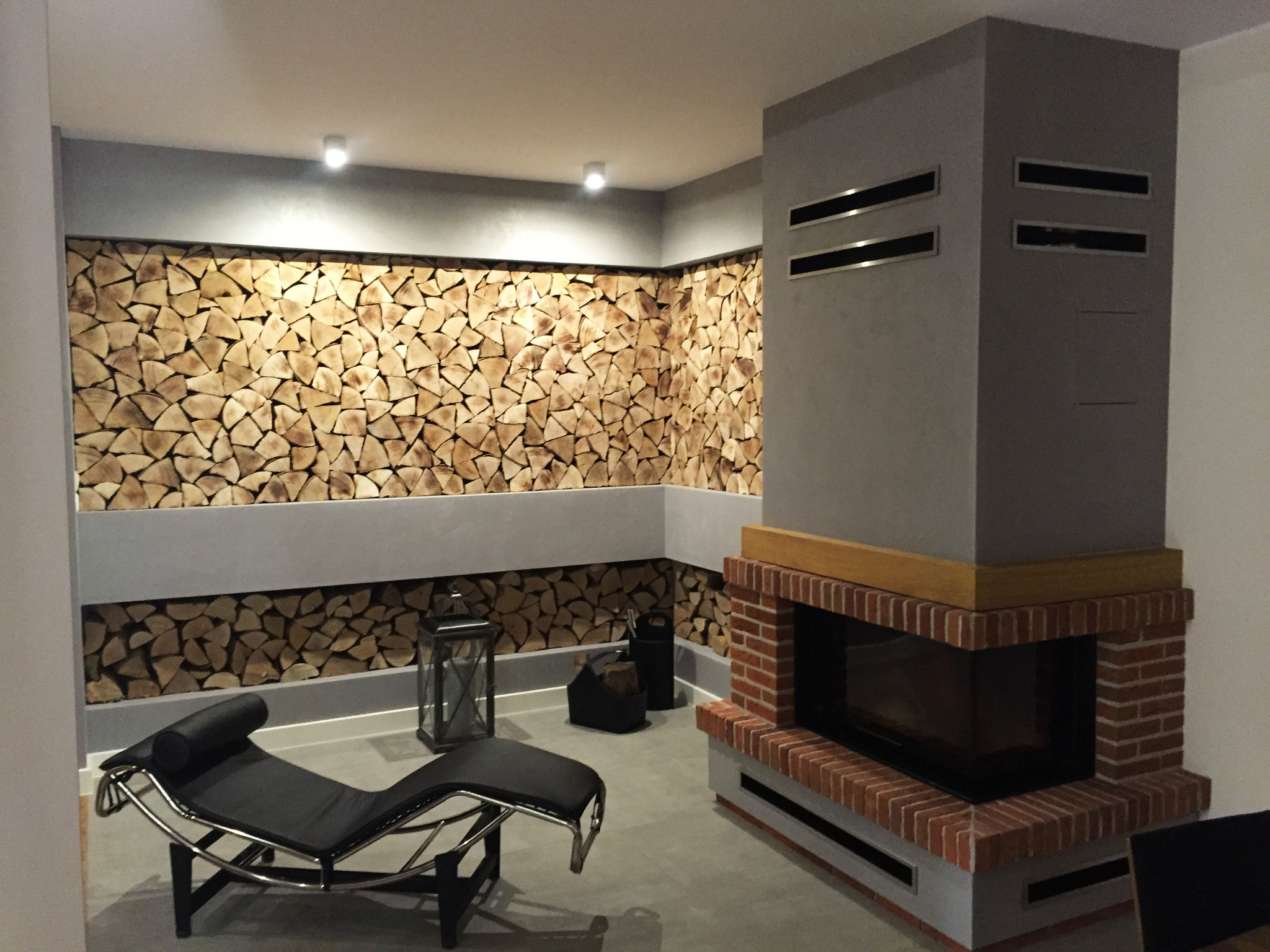 Nowoczesne wnętrze z kominkiem obudowanym cegłą i ścianą dekoracyjną z drewna opałowego, uzupełnione czarnym leżakiem i lampionem.