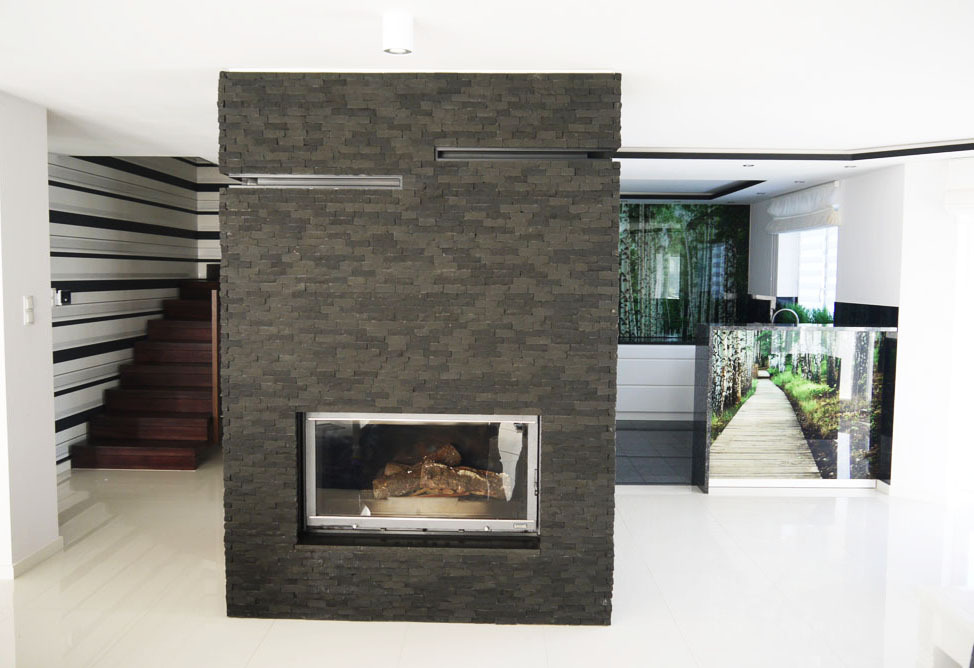 Nowoczesny salon z kominkiem obłożonym szarą, kamienną okładziną, widoczne schody z drewnianymi stopniami i fototapeta z motywem leśnej ścieżki w tle.