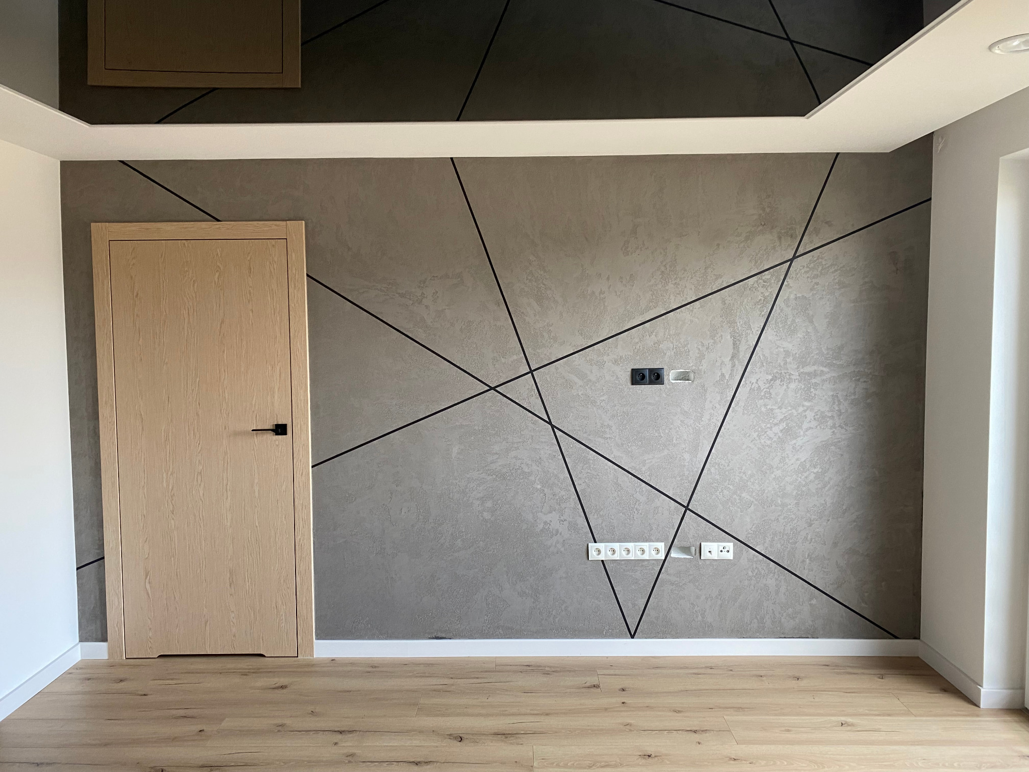 Szpachlowana ściana w szarości z geometrycznym wzorem z czarnych linii, jasne drewniane drzwi z czarną klamką, parkiet w odcieniach beżu, białe listwy przypodłogowe i sufitowe.