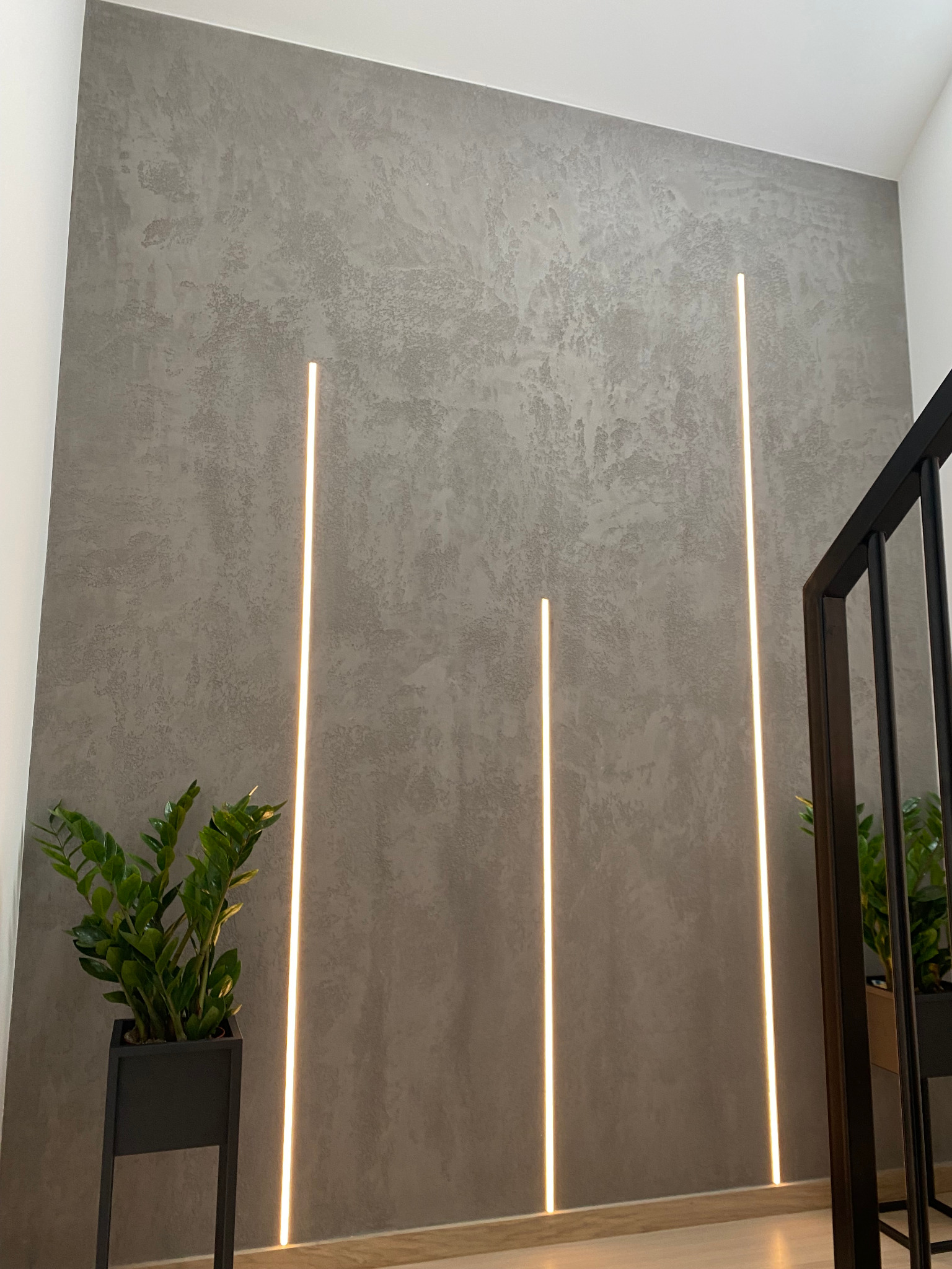 Ściana z szarą, dekoracyjną fakturą i trzema pionowymi paskami ledowego oświetlenia, z rośliną doniczkową w czarnej, metalowej donicy po lewej stronie i fragmentem czarnej balustrady po prawej.