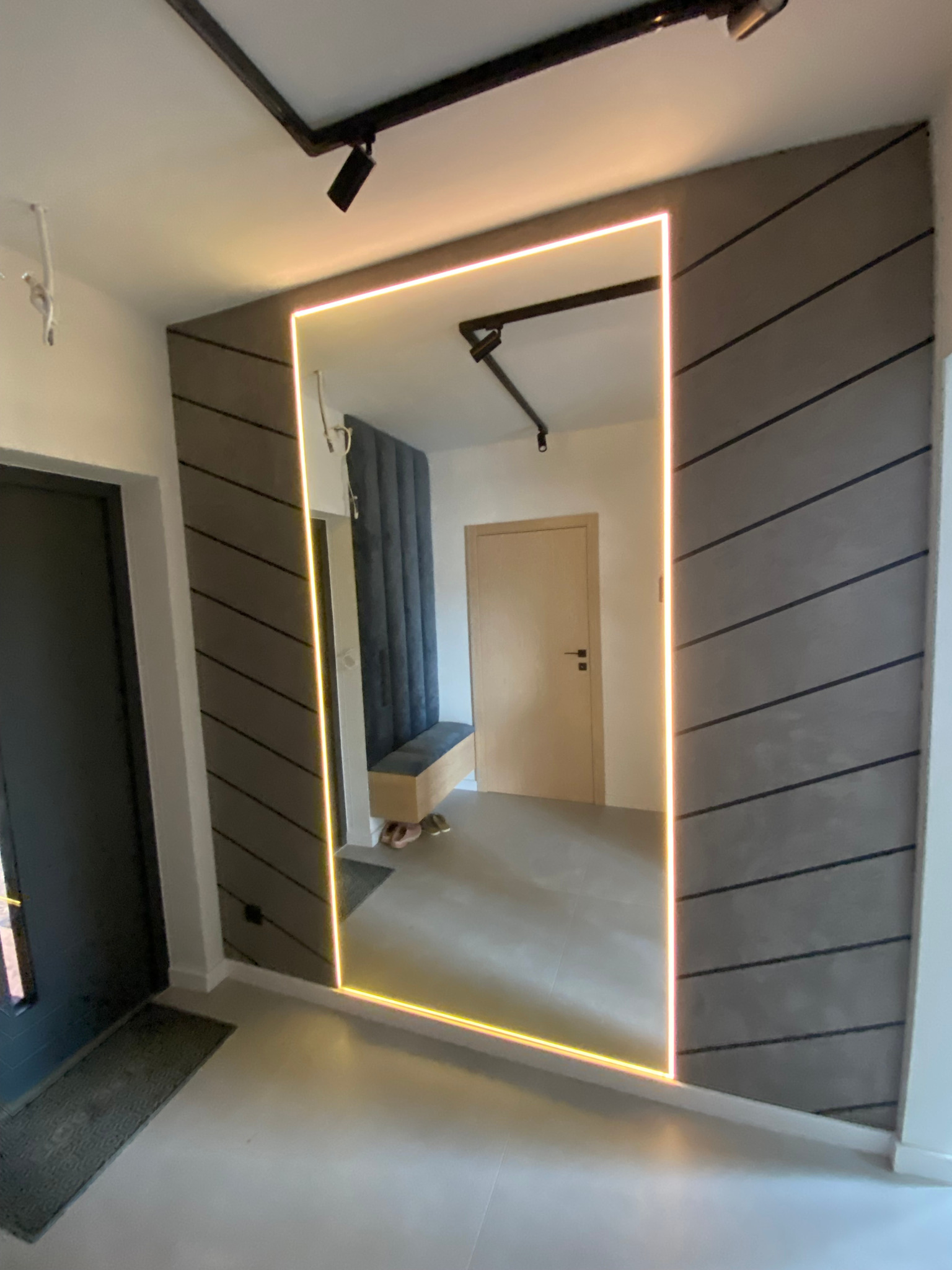 Nowoczesne lustro z podświetleniem LED w przedpokoju z szarą ścianą w skośne pasy, odbijające wnętrze z tapicerowanym panelem ściennym i siedziskiem.