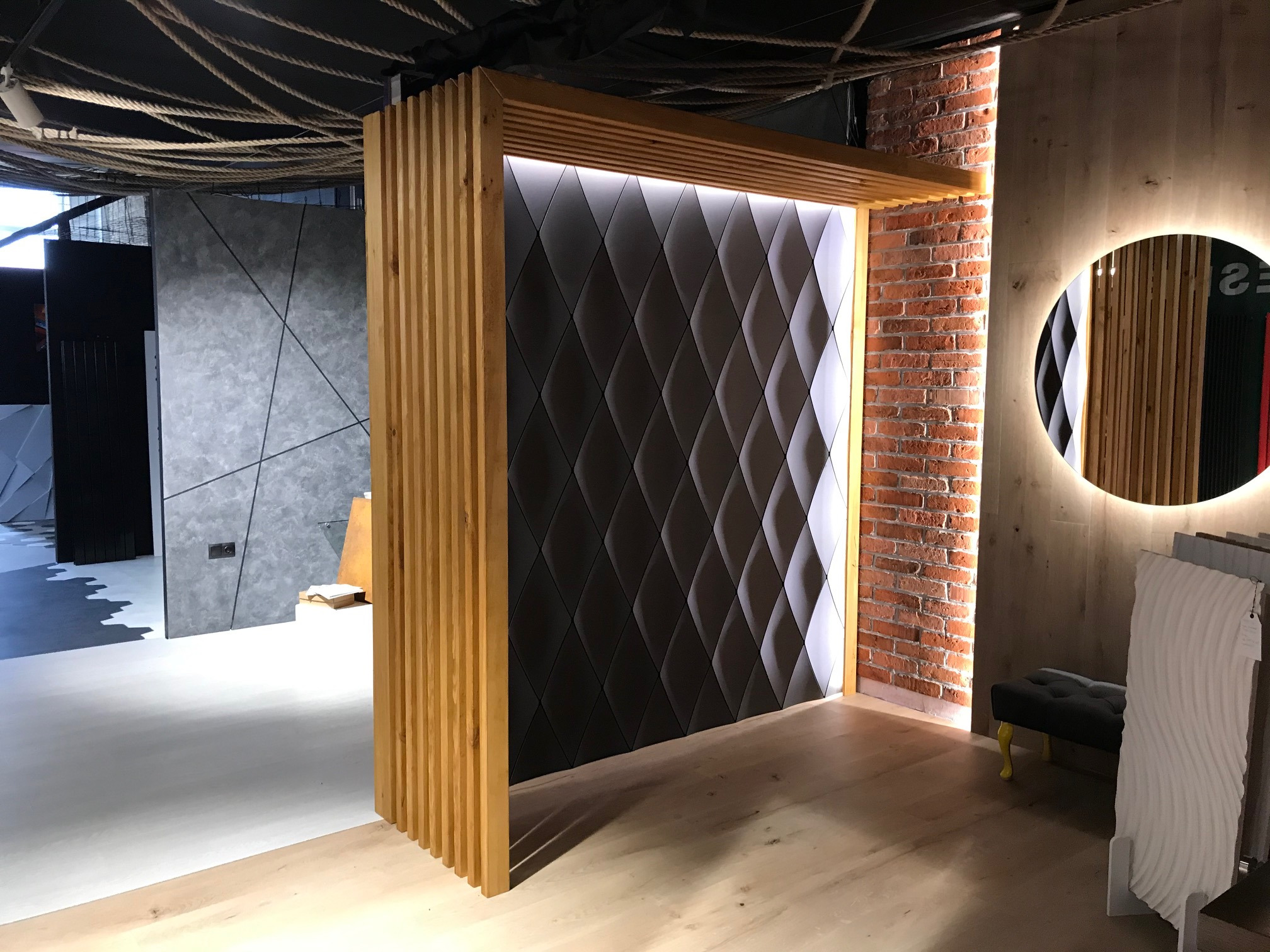 Nowoczesne wnętrze: panel ścienny z geometrycznym wzorem rombów podświetlony od góry, drewniana konstrukcja z pionowych listew, fragment ściany z czerwonej cegły oraz okrągłe lustro...