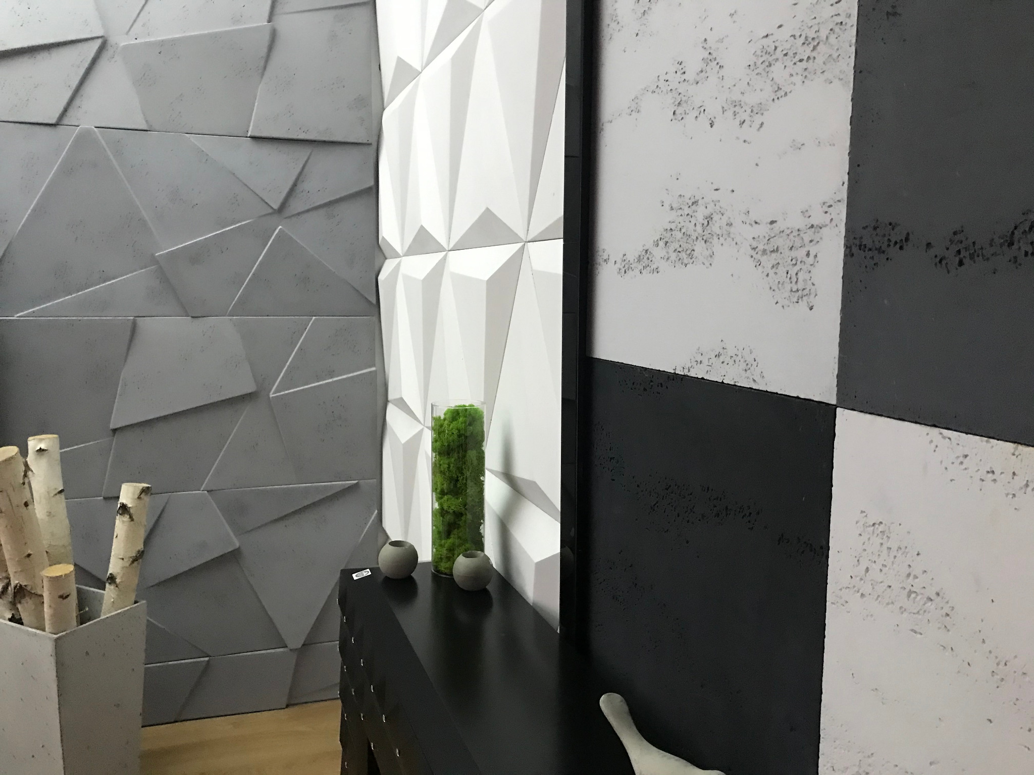 Nowoczesne wnętrze z trójwymiarowymi panelami ściennymi w odcieniach szarości i bieli, uzupełnione czarną komodą z dekoracjami i wazonem z zielonym mchem.